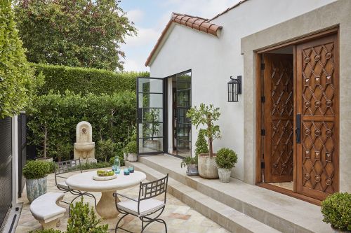 Exterior of a Mediterranean-style house with a stone patio, garden, and an ornamental fountain surrounded by lush greenery.