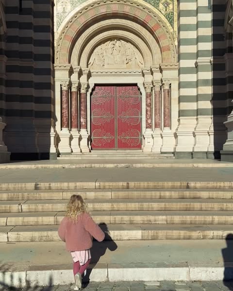 A young girl with blonde curly hair walks up stone stairs towards the ornate doors of a striped stone building on a sunny day.