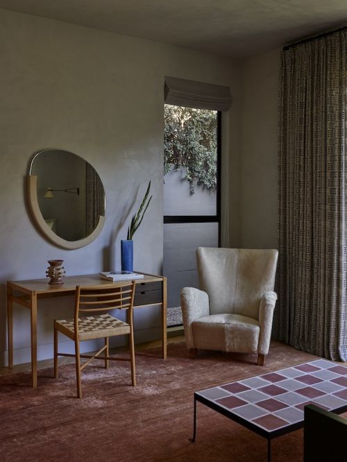 Living room interior with a wingback chair, desk with a mirror, patterned curtains, and tiled coffee table on a reddish-brown rug.