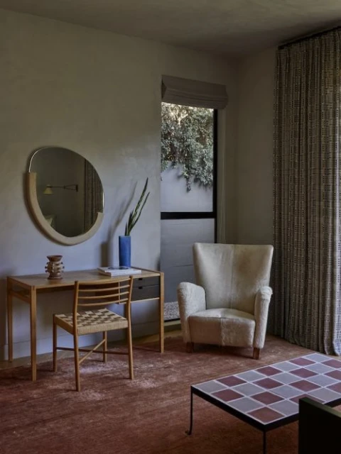 Living room interior with a wingback chair, desk with a mirror, patterned curtains, and tiled coffee table on a reddish-brown rug.