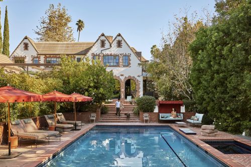Exterior view of a Tudor-style home with a swimming pool, lounge chairs, and patio furniture in a backyard with mature trees and blue sky.