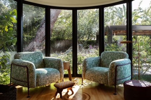 Living room interior featuring two patterned armchairs and a small ottoman, complemented by a large window view of lush greenery.
