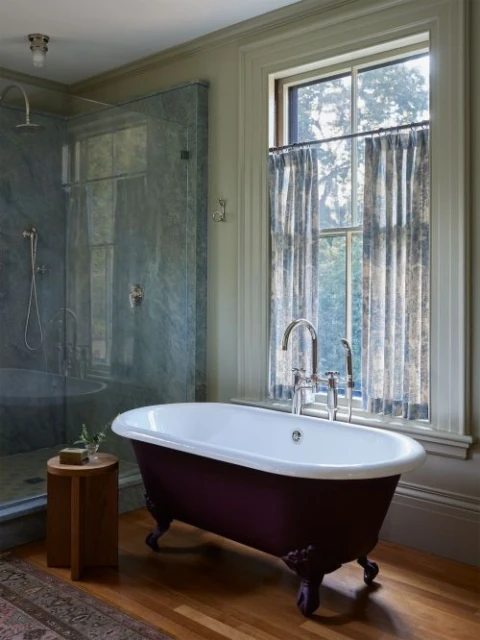 Freestanding bathtub with claw feet next to a window in a bathroom, a stool, and a glass shower area, enhancing the bathroom's appeal.
