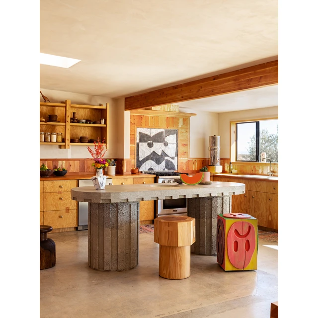 Eclectic kitchen interior with custom wood cabinetry, textured concrete island, artistic decor, and natural light from a window and skylight.