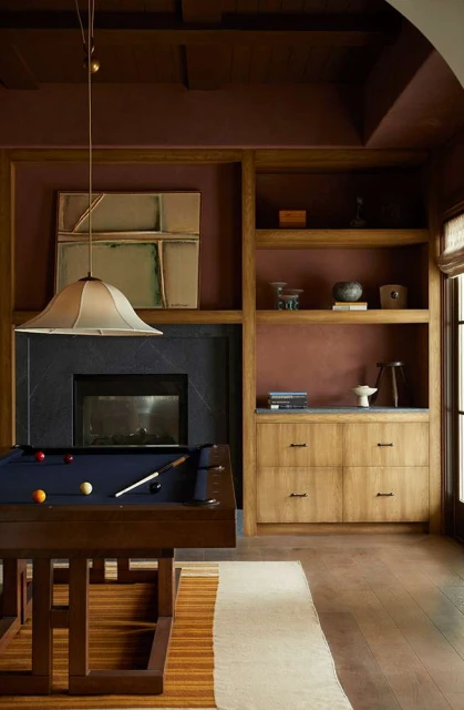 Indoor shot of a billiards room featuring a pool table, dark fireplace, built-in shelving, and earth-toned color palette.