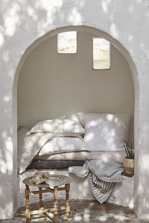 Cozy nook with white pillows, stone decor, and a small table inside a stucco archway bathed in soft, dappled sunlight and shadows.