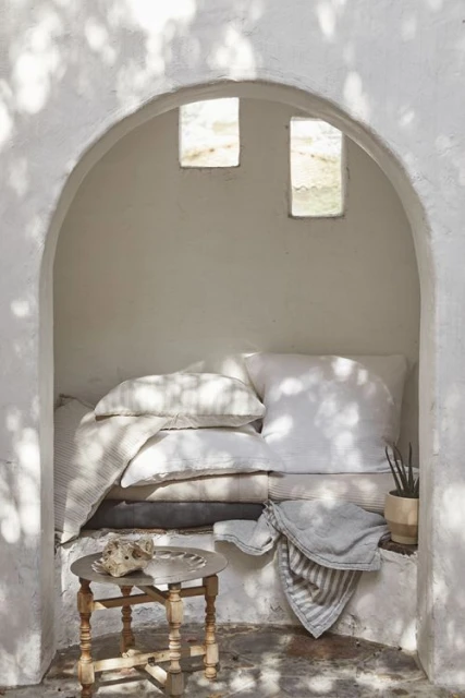Cozy nook with white pillows, stone decor, and a small table inside a stucco archway bathed in soft, dappled sunlight and shadows.