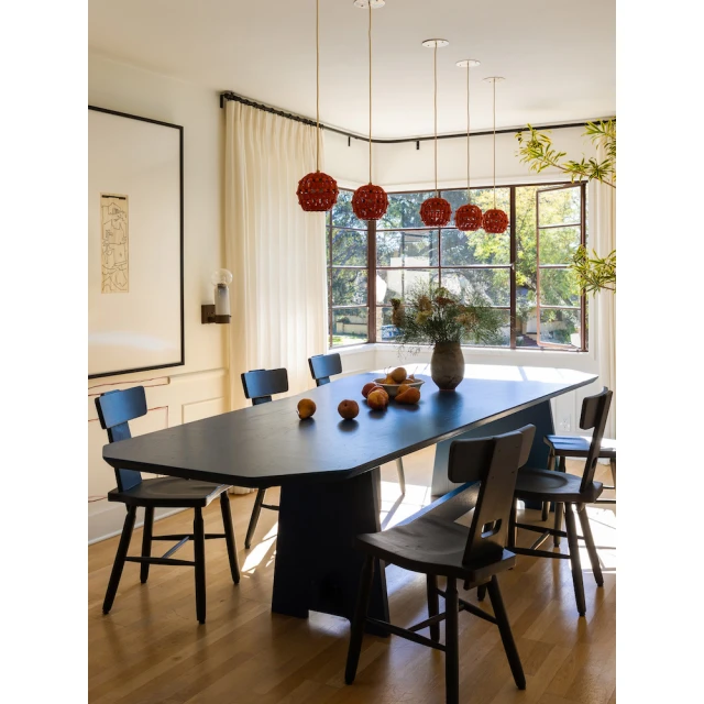 Dining room interior: a dark wood dining table, chairs, hanging red pendant lights, fruit, flowers, and a large window with natural light.
