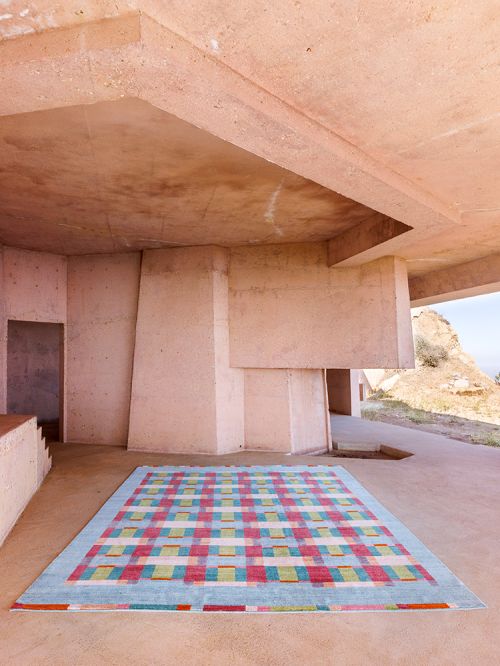 Indoor setting featuring a vibrant, patterned rug under a concrete structure with natural light filtering in from an exterior view.