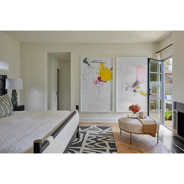 Bright bedroom interior featuring two framed abstract paintings, an upholstered ottoman with flowers, and a contemporary patterned rug.