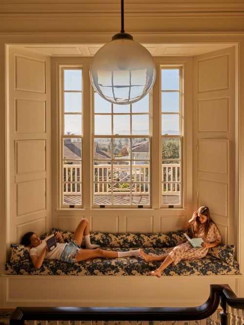 Two young women are reading in a brightly lit bay window seat in a home, with a view of the outside landscape and a hanging light.