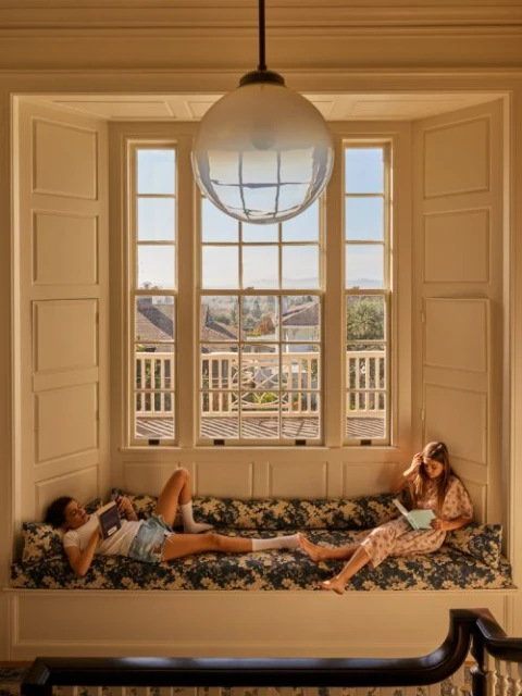 Two young women are reading in a brightly lit bay window seat in a home, with a view of the outside landscape and a hanging light.