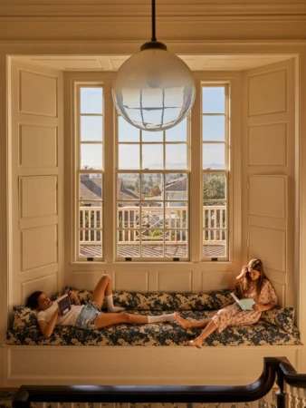 Two young women are reading in a brightly lit bay window seat in a home, with a view of the outside landscape and a hanging light.