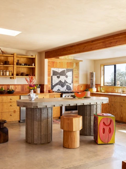 Eclectic kitchen interior featuring a large concrete island, wooden stools, and unique decorative elements in a modern, artistic home.