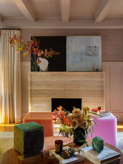 Modern living room interior features a coffee table with books and flowers, cube ottomans, and artwork over a travertine fireplace.