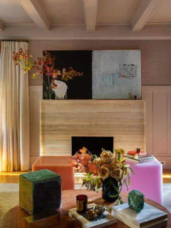 Modern living room interior features a coffee table with books and flowers, cube ottomans, and artwork over a travertine fireplace.