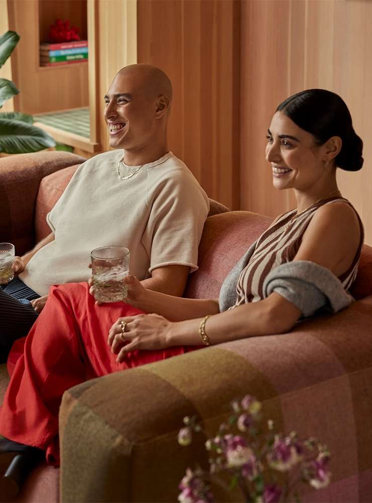 Smiling couple seated on a sofa in a brightly lit living room with modern decor, indoor plants, and textured walls creating a cozy scene.