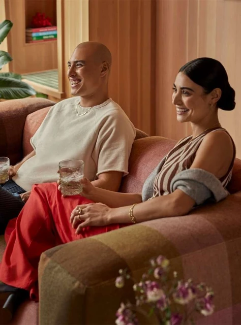 Smiling couple seated on a sofa in a brightly lit living room with modern decor, indoor plants, and textured walls creating a cozy scene.