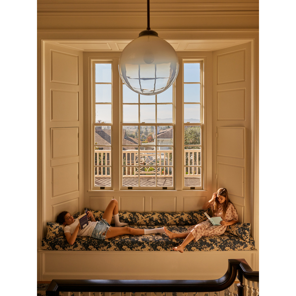 Two young women relax and read on a window seat in a sunlit room with white walls and a view of distant houses and trees.