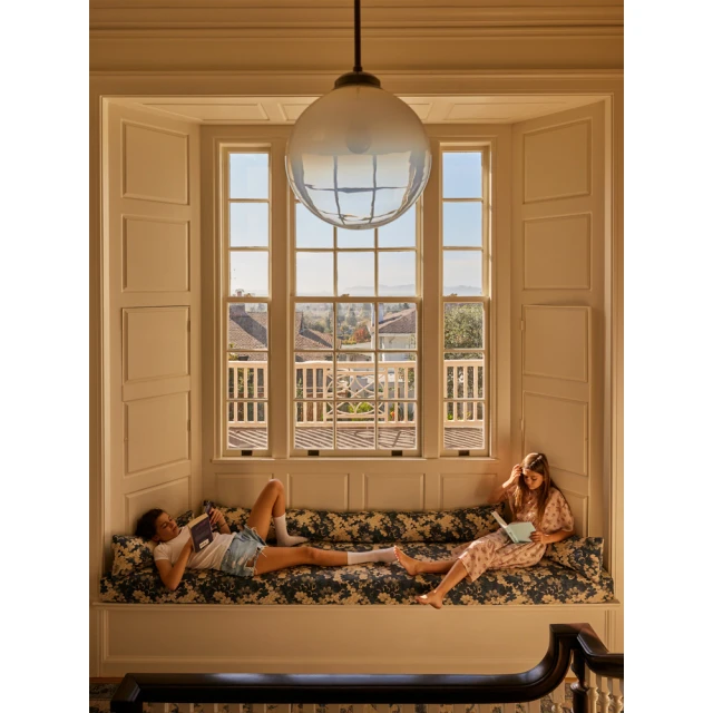Two young women relax and read on a window seat in a sunlit room with white walls and a view of distant houses and trees.