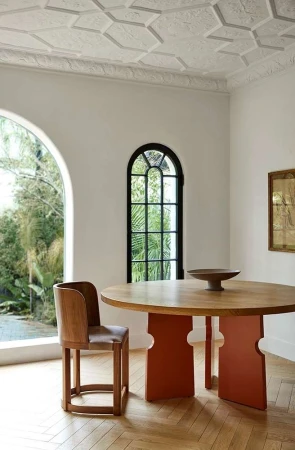 Interior with a light wood round dining table with burnt orange base, surrounded by a wooden chair and a room with an arch window.
