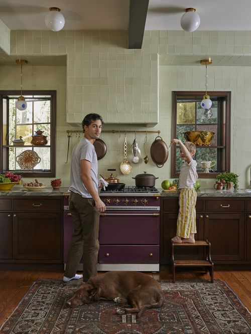 Man and child in a kitchen with a dog resting on a rug, featuring a vintage stove, hanging pots, and natural light from windows.