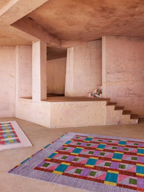 Interior with a geometric multicolored rug on a concrete floor, concrete walls, and stairs in an open indoor space. 