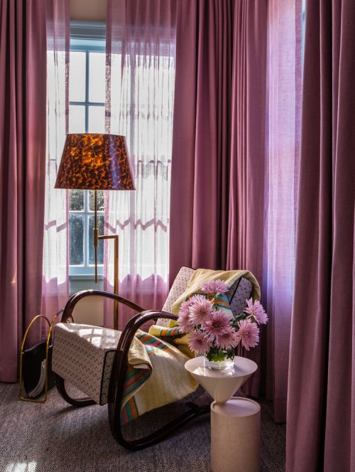 Interior view of a mauve living room with an armchair, side table with flowers, patterned lamp, and window with pink curtains.