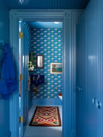 Blue-toned powder room with patterned blue wallpaper, blue sink, wood-paneled walls, framed art, and a patterned area rug on the floor.