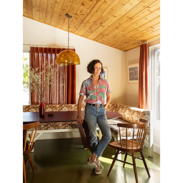 Indoor portrait of a woman in jeans and a patterned shirt, standing in a home dining room with wood furniture and an amber pendant light.