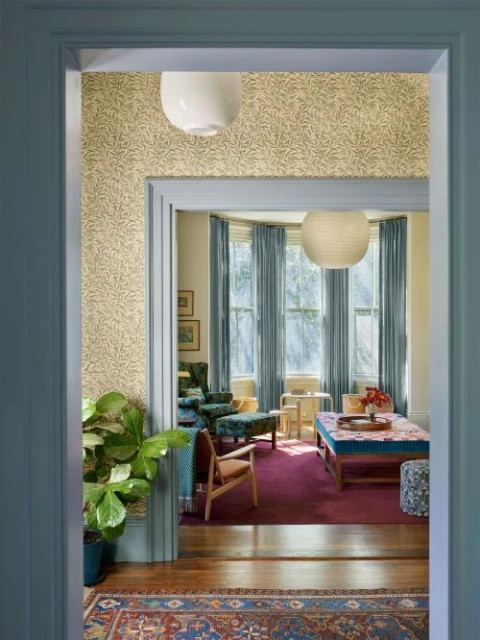 Cozy living room interior with vintage furniture, patterned wallpaper, and natural light streaming through the windows. 