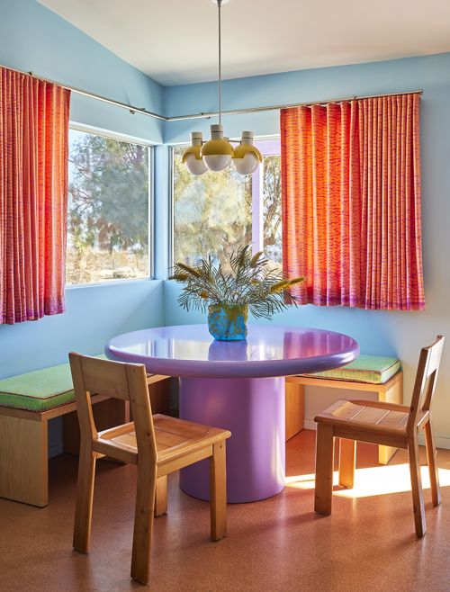 Bright and colourful dining space with round table, wood chairs, orange curtains, light blue walls, and a three-bulb pendant light.