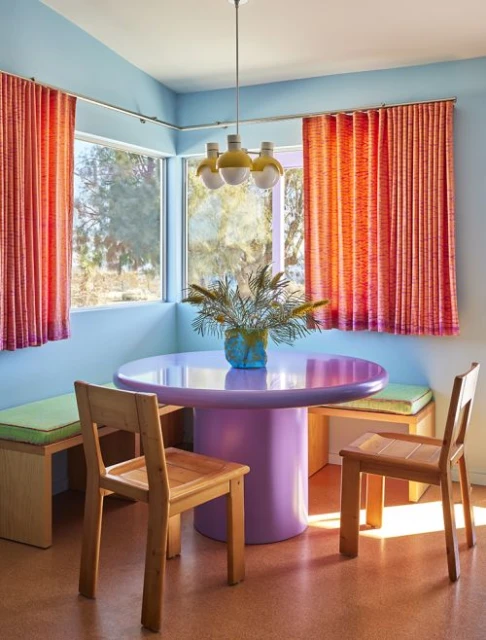 Bright and colourful dining space with round table, wood chairs, orange curtains, light blue walls, and a three-bulb pendant light.