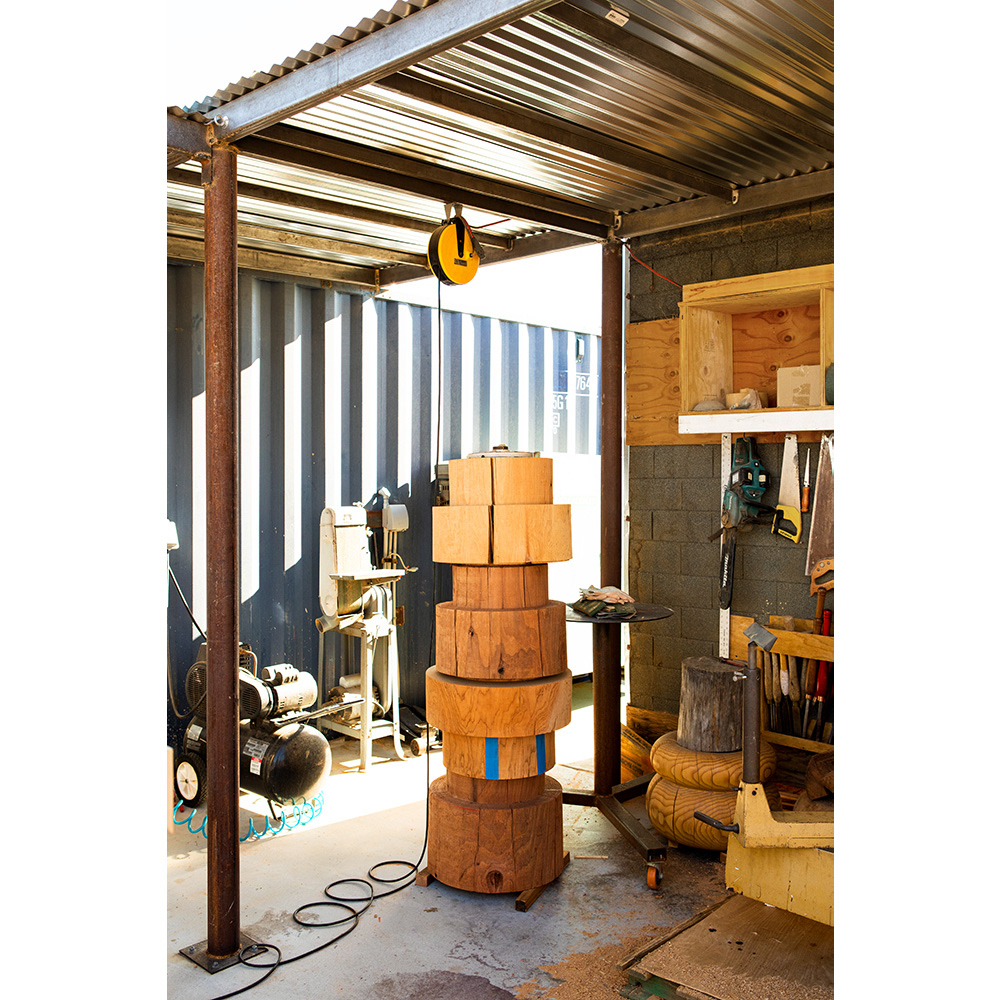 Indoor workspace featuring a stack of wooden blocks, tools, and equipment beneath a corrugated metal roof, showcasing craftsmanship.