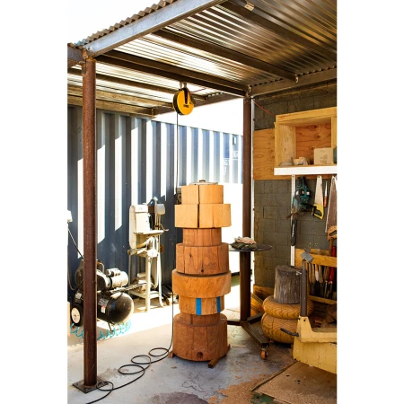 Indoor workspace featuring a stack of wooden blocks, tools, and equipment beneath a corrugated metal roof, showcasing craftsmanship.