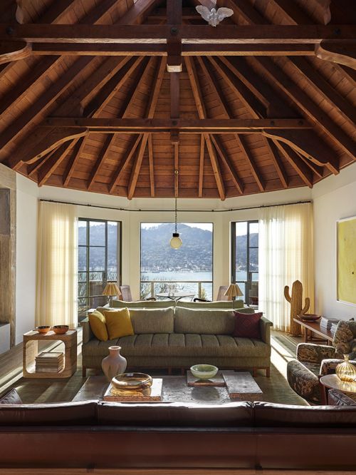 Living room interior with a large sofa, wooden coffee table, floor lamp, and a view of a lake and mountains through large windows.