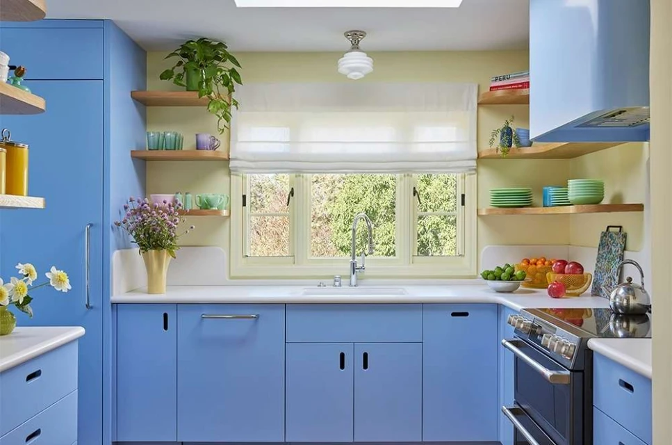 Bright kitchen interior with blue cabinets, open wooden shelves displaying dishware, flowers in vases, and stainless steel appliances.