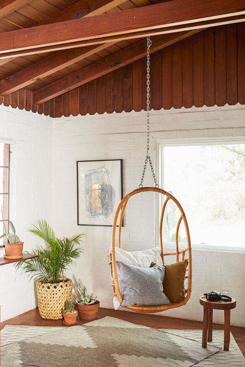 Interior corner features a hanging rattan chair, patterned rug, and potted plants, illuminated by natural light from nearby windows.
