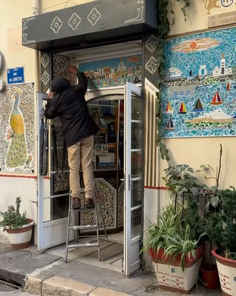 Adult stands on ladder in front of a shop entrance, working on a mosaic of a coastal town with boats. Plants in pots flank the entrance.