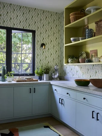 Stylish kitchen interior featuring light blue cabinets, wood floors, and patterned wallpaper with natural light streaming through a window.