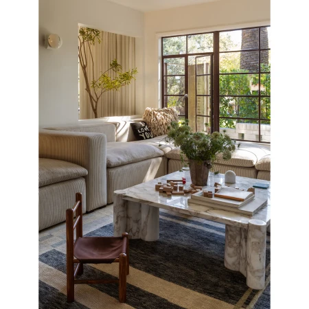 Living room interior with a sectional sofa, marble coffee table with objects, a patterned rug, and a window offering natural light.