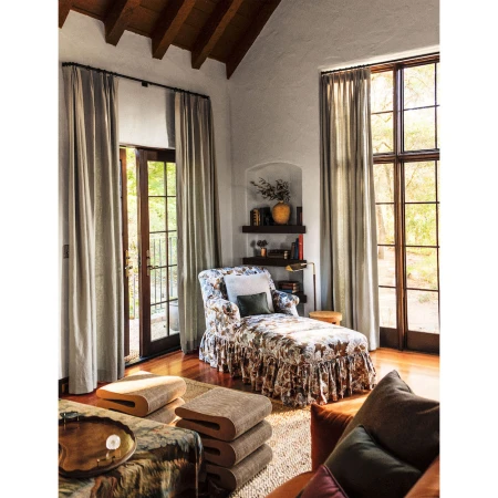Interior living space featuring a floral chaise lounge, wooden floors, and tall windows with light curtains.