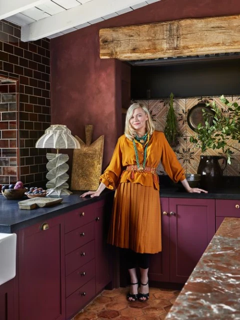 Woman poses in a kitchen with maroon cabinets, copper accents, and rustic wooden beams, illuminated by unique lamps and natural light.