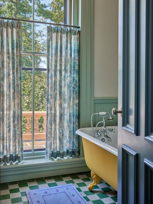 Bathroom interior with yellow bathtub, green and white tiled floor, blue rug, and window with printed curtains overlooking trees.