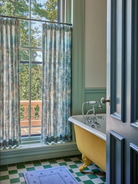 Bathroom interior with yellow bathtub, green and white tiled floor, blue rug, and window with printed curtains overlooking trees.