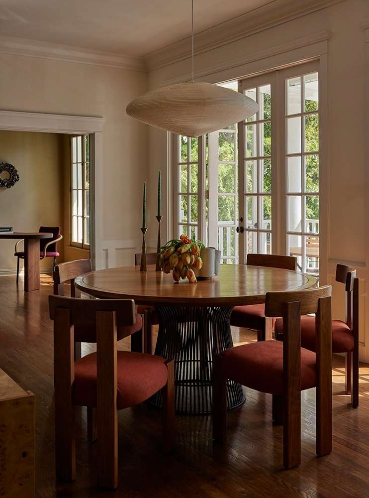 Round wooden dining table with six chairs, pendant light, and fruit centerpiece in a bright, neutral-toned dining room setting.