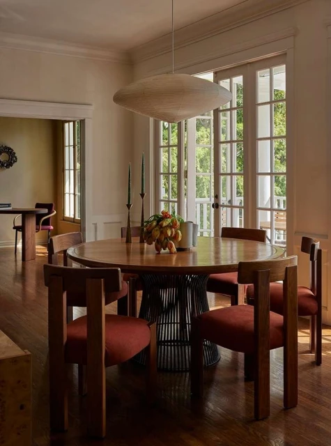 Round wooden dining table with six chairs, pendant light, and fruit centerpiece in a bright, neutral-toned dining room setting.