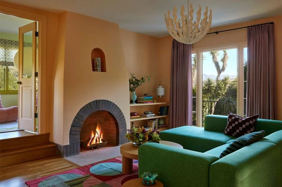 Living room interior with green sectional sofa, fireplace, and view to outdoor garden. Warm lighting and cozy design in home setting.