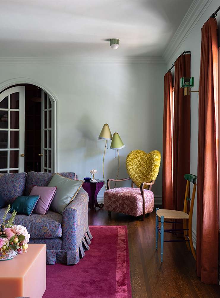 Eclectic living room with floral sofa, yellow heart chair, magenta rug, lamps, curtains, and decorative accessories inside a home.