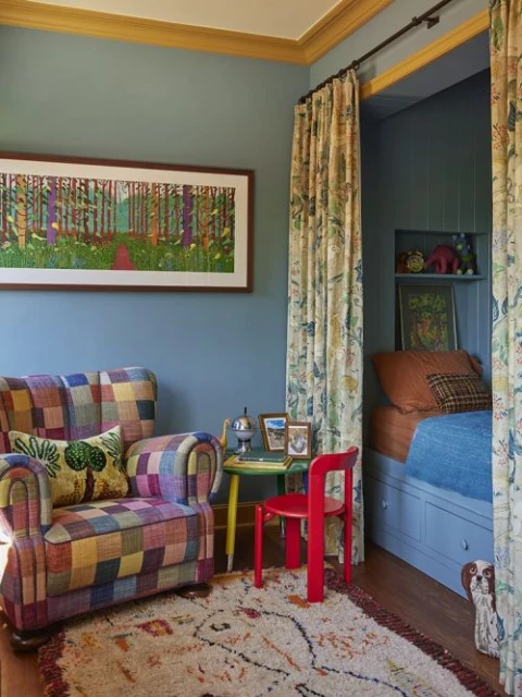 Interior of a child's bedroom with a bed nook, patchwork armchair, colorful painting, patterned rug, and a small table and chair.